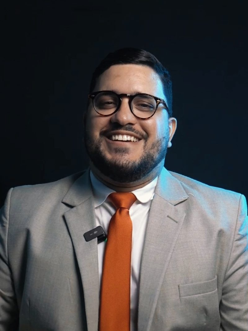 Lucas Açucena, advogado, de terno claro e gravata laranja, sorrindo em fundo escuro